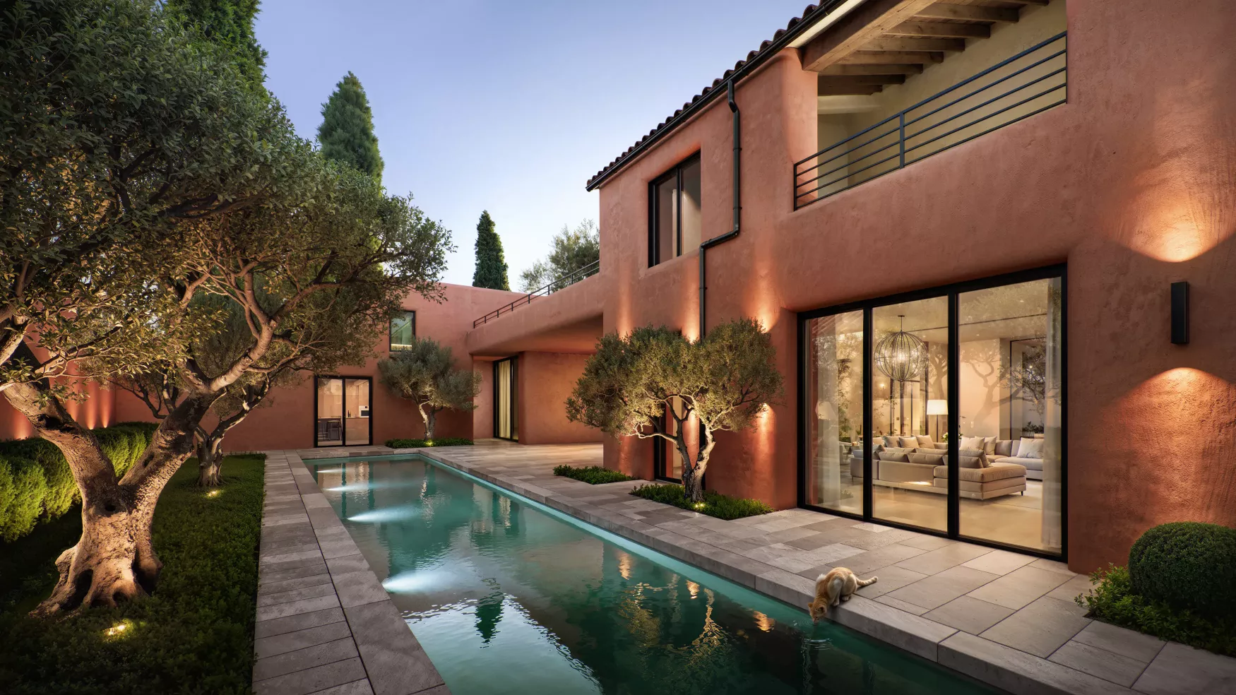 Modern villa with a narrow pool, surrounded by trees and lit warmly at dusk, with a cat drinking water at the pool's edge.