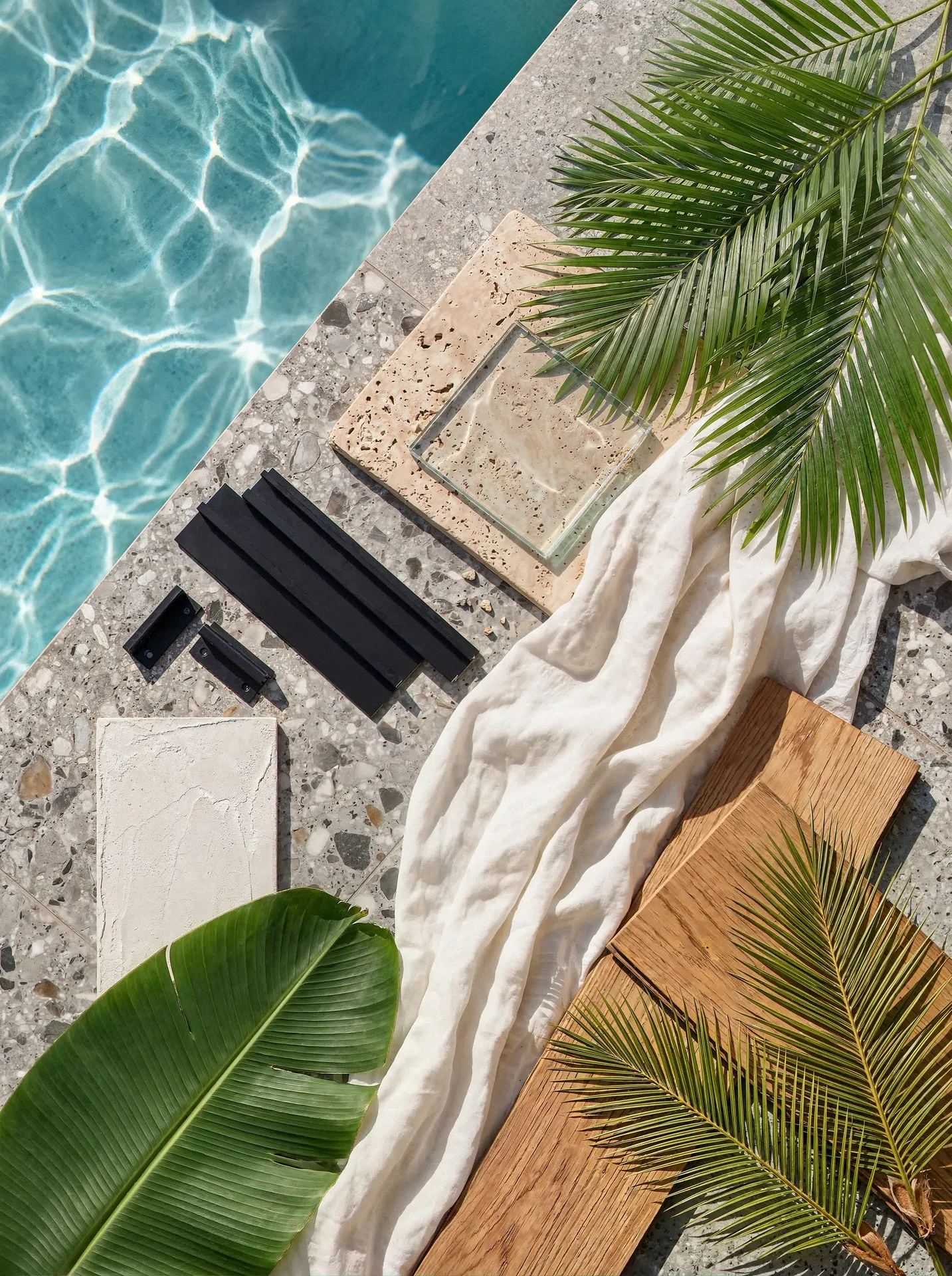 Architectural material mood board flat lay featuring terrazzo, warm wood planks, travertine stone, black metal, and white linen beside a sunlit swimming pool.