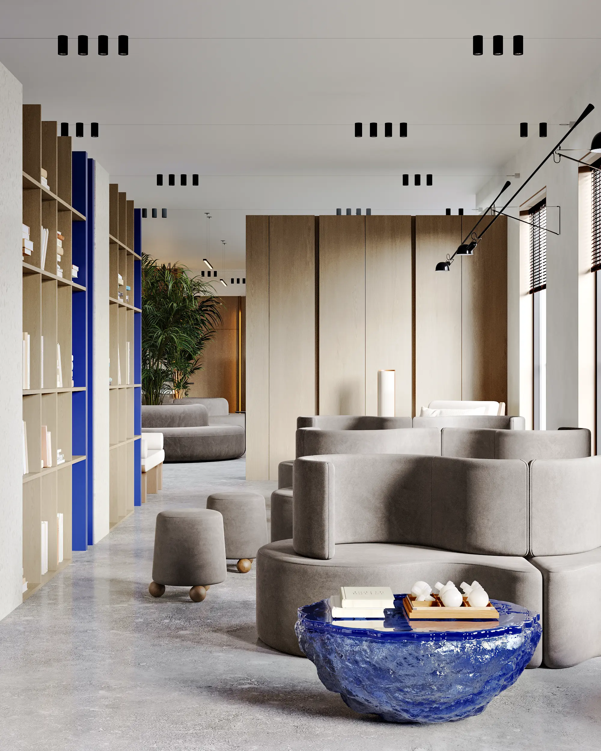 Modern lounge with gray curved sofas, small rounded stools, tall wooden shelves, and a unique blue textured coffee table topped with books and a tray.